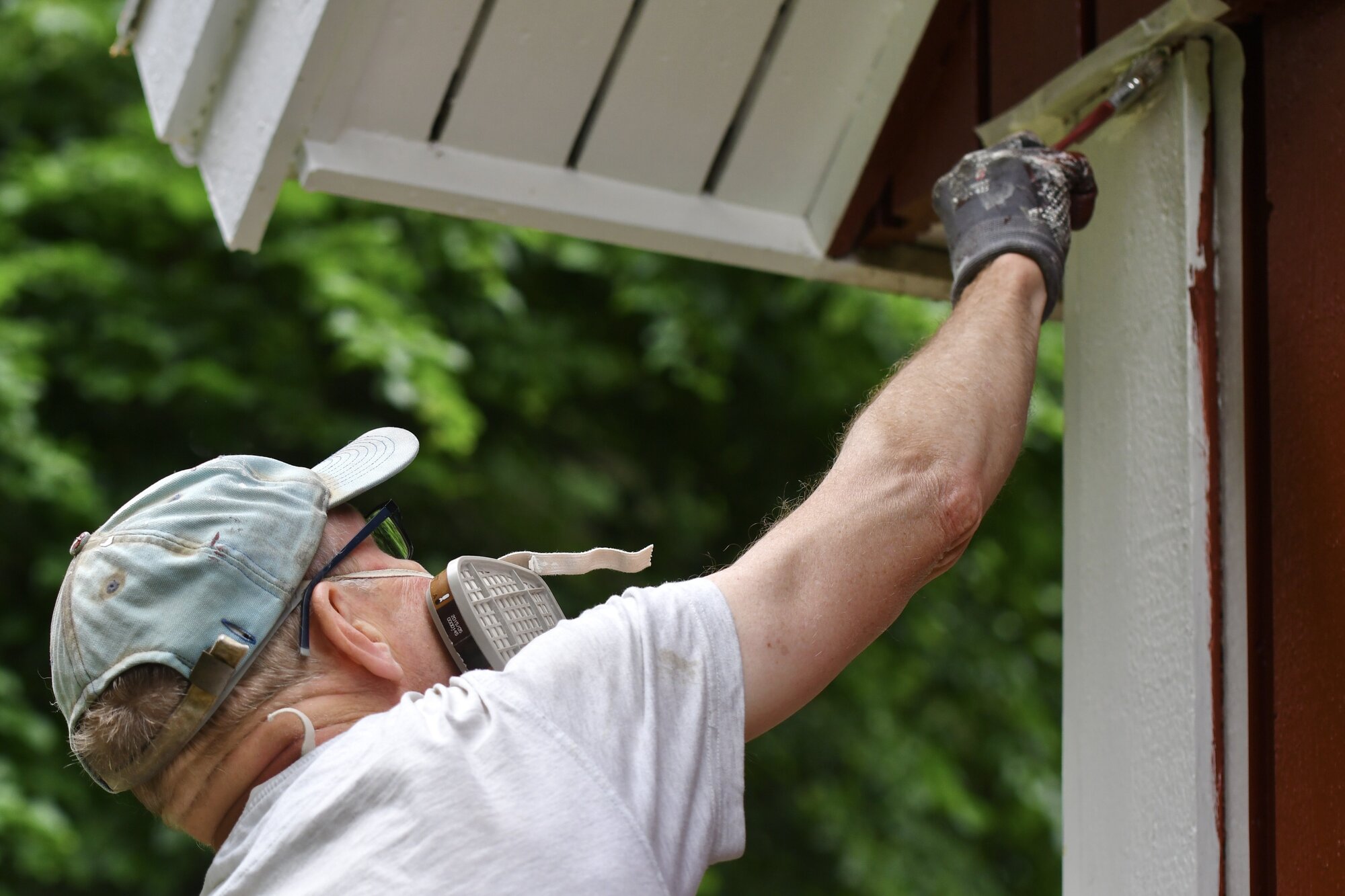 A person wearing a backwards baseball cap, safety glasses, and a respirator mask is painting the outside of a building. They are reaching up with a paintbrush to apply white paint to a vertical wooden board. The person is wearing a white shirt and disposable gloves. The background is blurred greenery.
