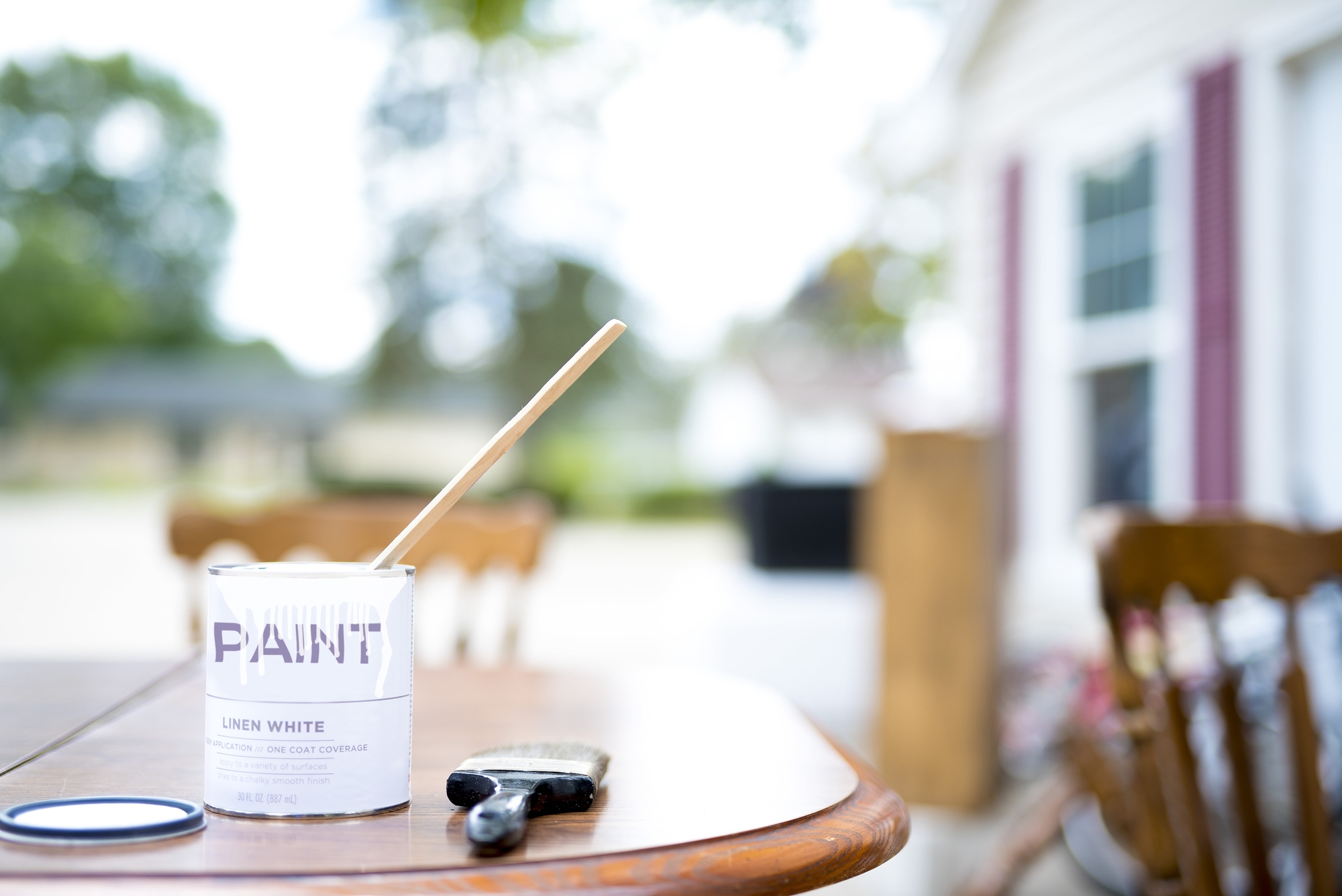 A can of "Linen White" paint sits on an outdoor table, with an open lid nearby. A wooden stick is inside the can, and a paintbrush rests on the table next to it. The background features blurred greenery and a portion of a house, suggesting a calm outdoor setting.