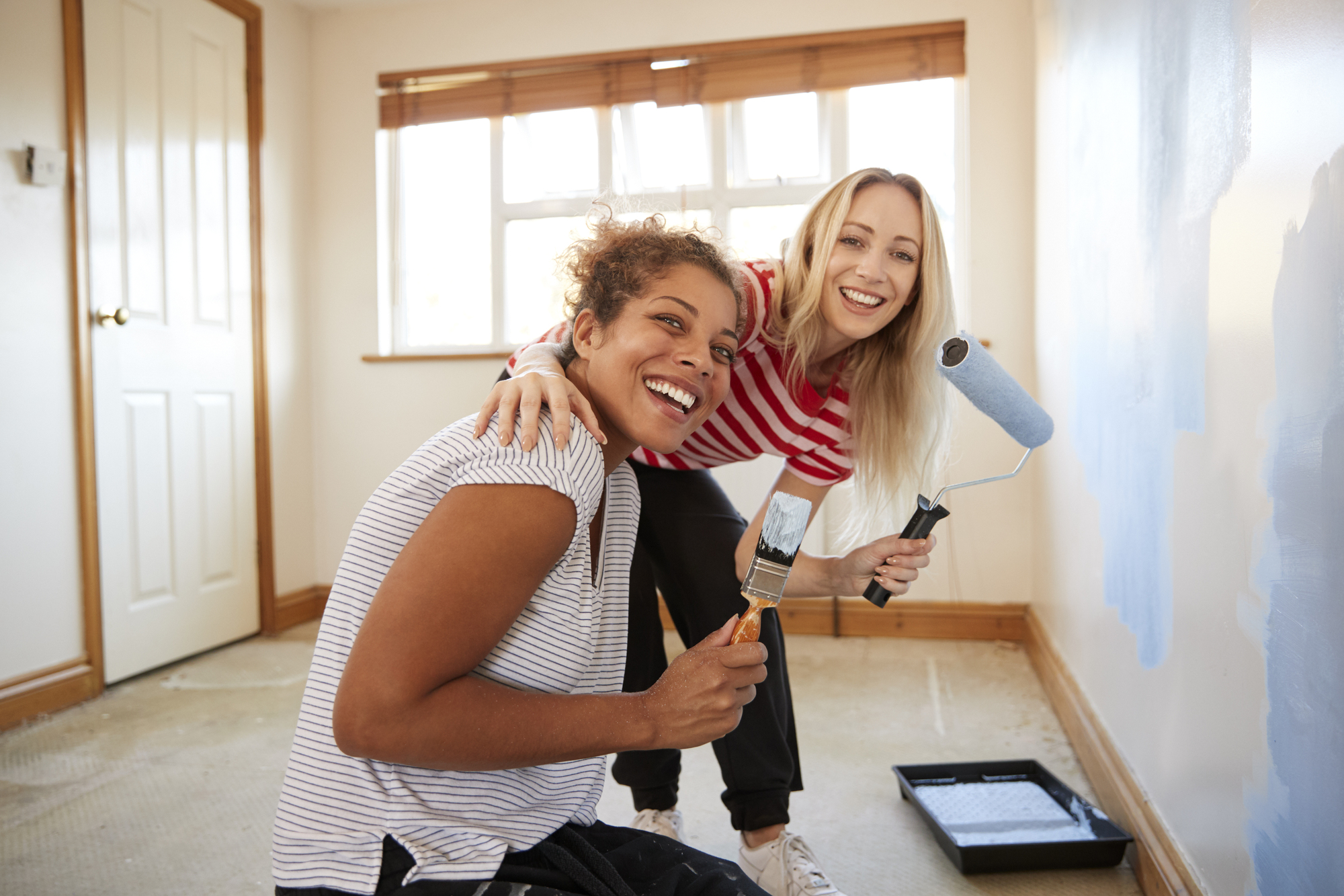 Two women are smiling while painting a wall in a room. One woman is holding a paintbrush, and the other has a roller. The wall they are painting is light blue. They are wearing casual clothes, and a paint tray is on the floor. Light is coming in through a window in the background.