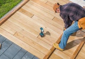 Caucasian Men in His 40s Building Wooden Deck on His Backyard Attaching Wood Board To Each Other Using Cordless Drivers and Screws. Construction Industry Theme.
